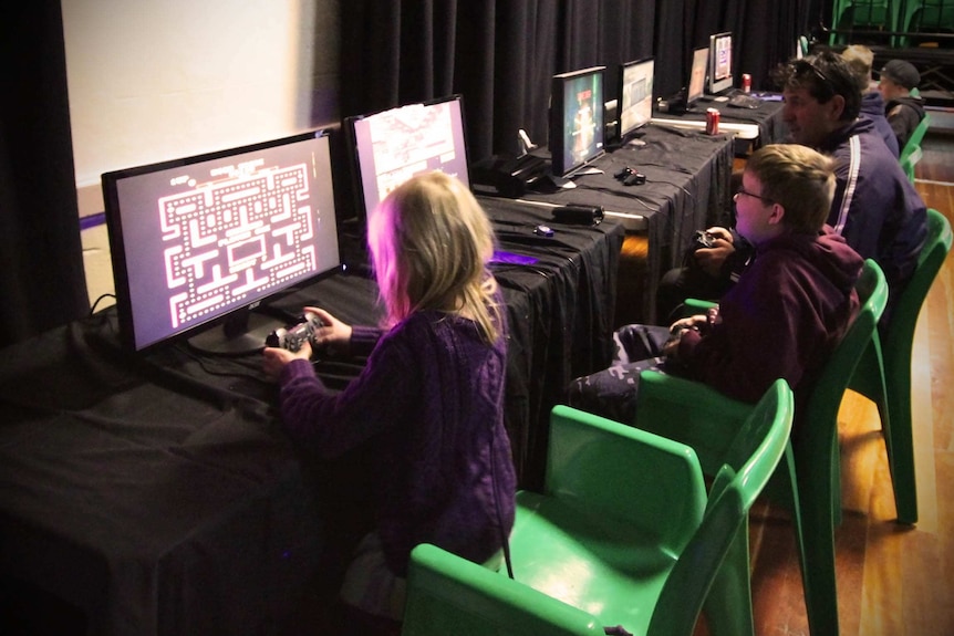 Two children and an adult playing video games on computers with other children in background