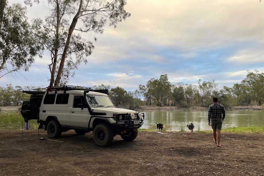 A 4WD parked by a river with a man and two dogs walking toward water.