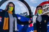 Laura Peel and Brendan Kerry wear masks and hold Australian flags, while standing in front of Olympic rings.
