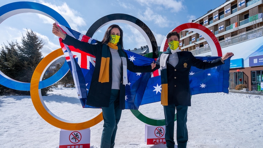 Laura Peel and Brendan Kerry wear masks and hold Australian flags, while standing in front of Olympic rings.