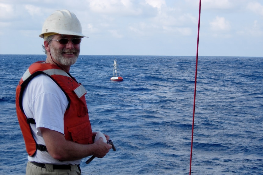 Michael McPhaden is standing on the deck of a boat. An ocean monitoring buoy is floating in the ocean in the background.