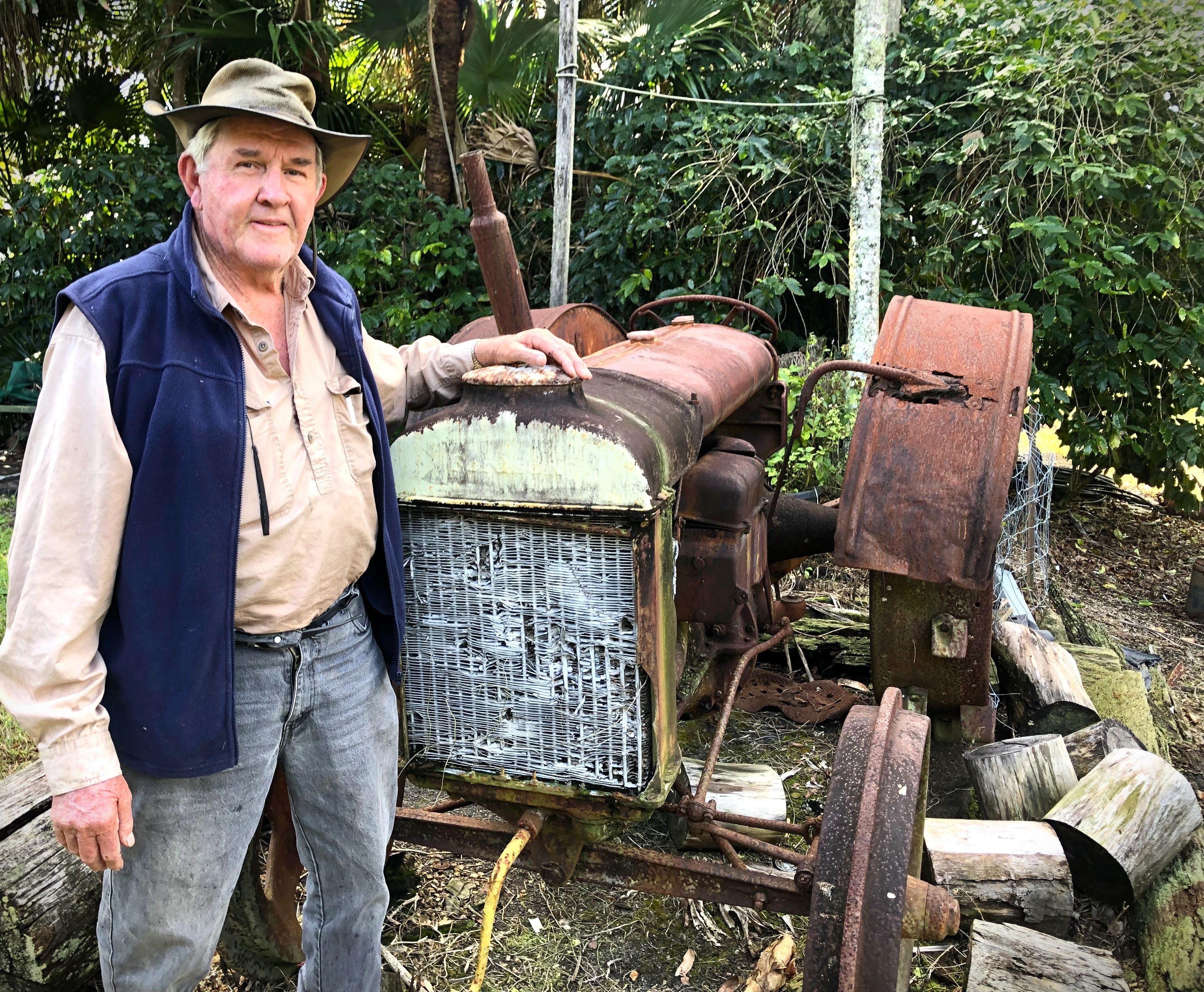 An elderly man leans against a rusty old tractor