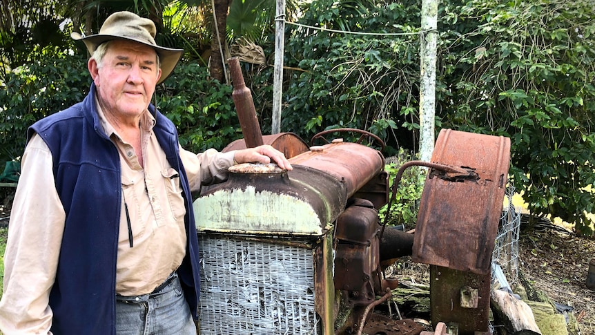 An elderly man leans against a rusty old tractor