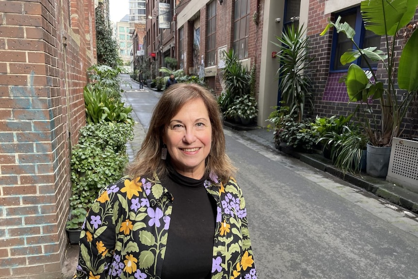A lady in a colourful coat standing in a laneway smiles at the camera