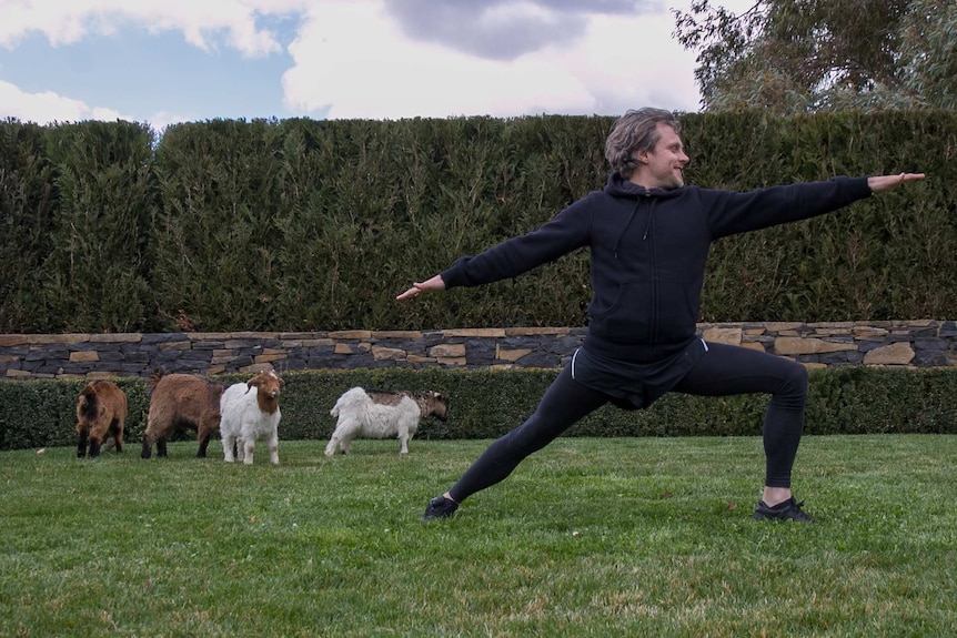 A man doing a yoga pose in a manicured garden with baby goats in the background