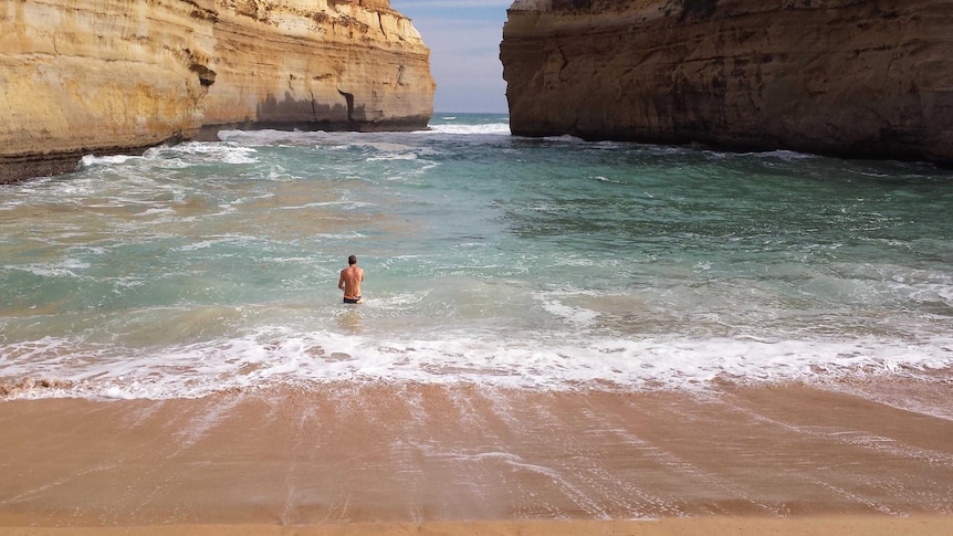 A swimmer walks into the small ocean waves breaking on the sand, with the entrance to the gorge in the distance.