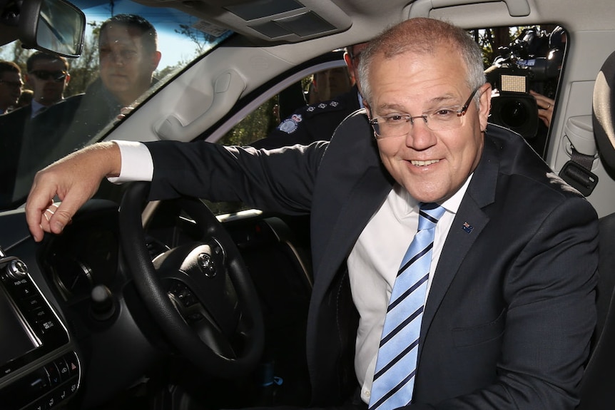 Scott Morrison grins, sitting in the driver's seat of a car with one arm on the steering wheel
