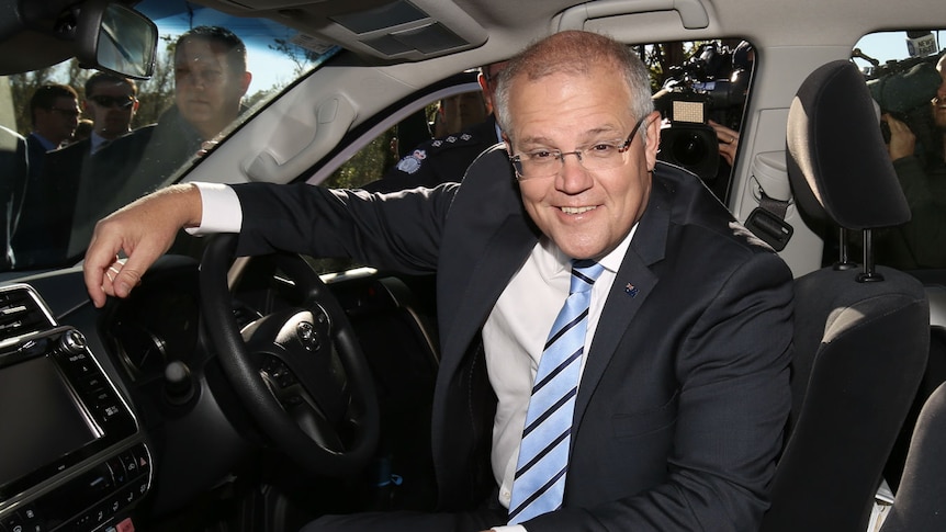 Scott Morrison grins, sitting in the driver's seat of a car with one arm on the steering wheel