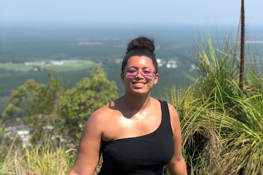 Yasmin wears a black one shoulder top and bike shops with purple tinted sunglasses while standing atop a mountain, smiling.
