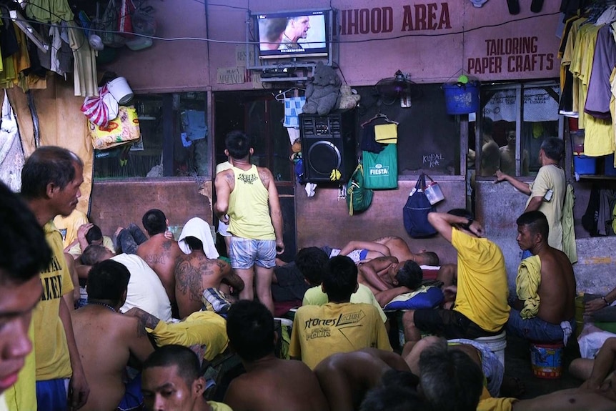 Prisoners sit on the floor in crowded room, watching a small flat screen TV mounted on the wall.