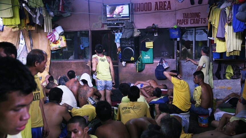 Prisoners sit on the floor in crowded room, watching a small flat screen TV mounted on the wall.