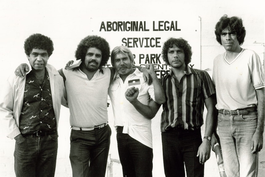 A black and white photograph of five men standing under an Aboriginal Legal Service sign.