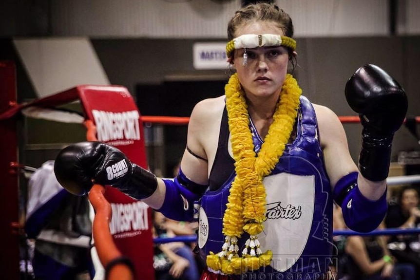 A woman wearing kickboxing gear stands in a boxing ring with a ring of yellow flowers around her neck.