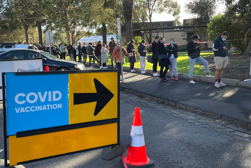A row of people along a footpath, next to a large sign surrounded by traffic cones reading 'COVID-19 VACCINATION'.