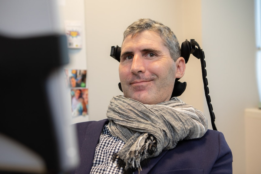 A man in a suit shirt and scarf, smiles, sitting in a wheelchair with neck and head support.