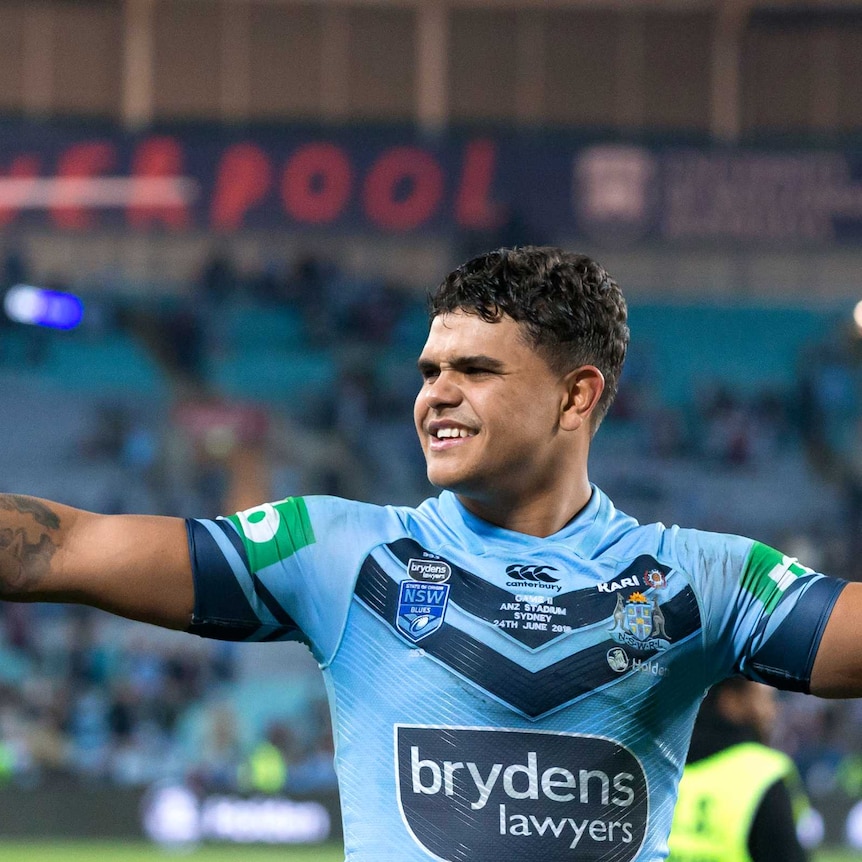 Latrell Mitchell holds his arms up as he looks to the left with a smile on his face. He wears a blue jersey with stadium in back