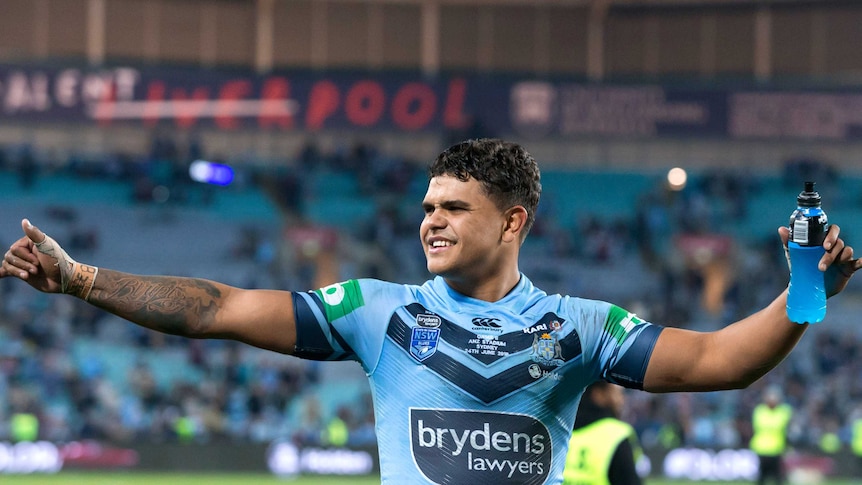 Latrell Mitchell holds his arms up as he looks to the left with a smile on his face. He wears a blue jersey with stadium in back
