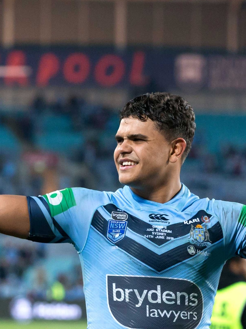 Latrell Mitchell holds his arms up as he looks to the left with a smile on his face. He wears a blue jersey with stadium in back