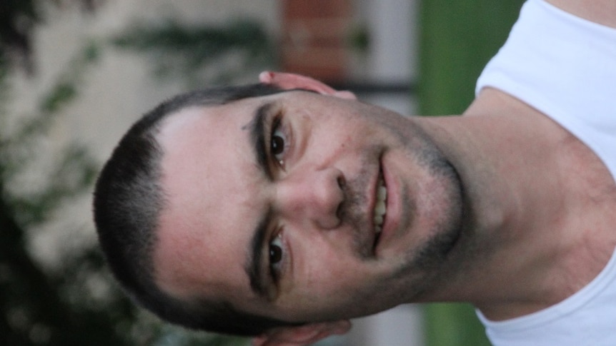 A young man in a white singlet is smiling at the camera