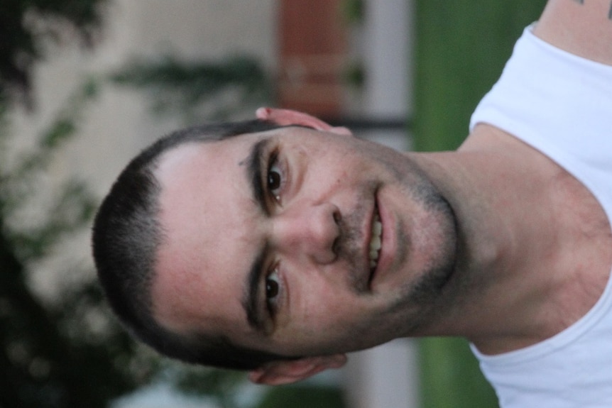 A young man in a white singlet is smiling at the camera