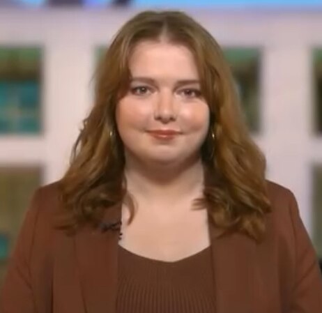 A white woman with red hair wearing a brown top and jacket in front of a picture of parliament house