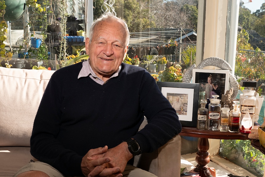 An eldelrly man pictured at home surrounded by plants and a side table with a row of very old medicine bottles.