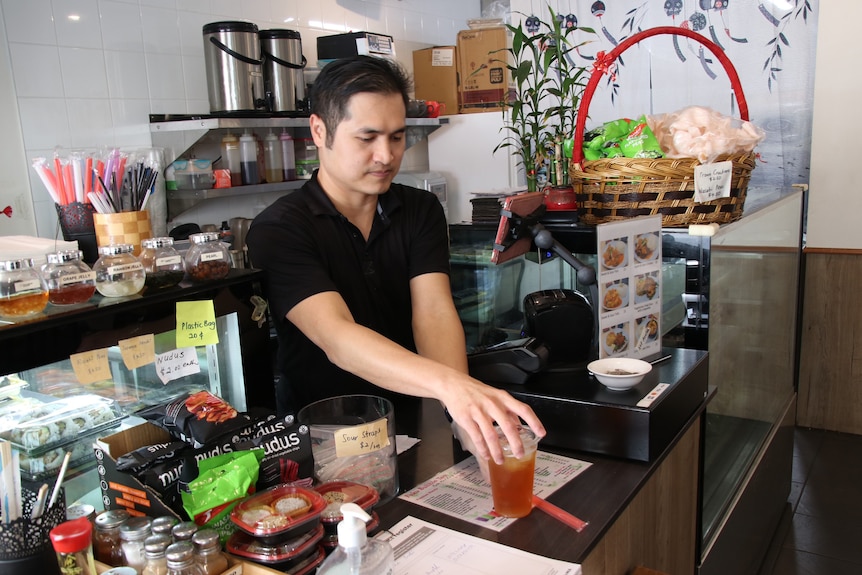 A man serves a drink in a plastic cup over a counter in a cafe.
