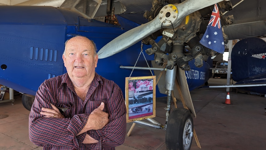 An older man in a checked shirt stands with his arms crossed in front of a light aeroplane replica