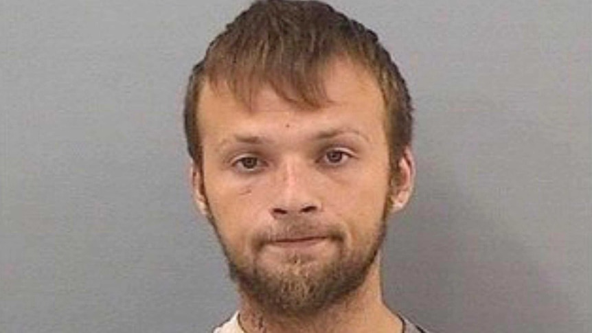 A police mugshot of a young man with light brown hair, brown eyes, a beard, small scar under his eye, in a grey striped shirt.