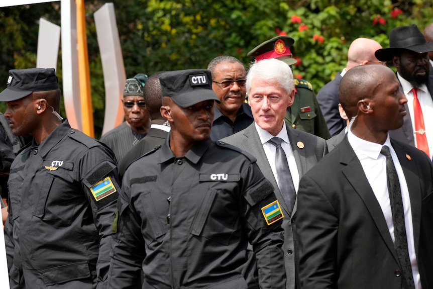 An older white man with white hair in a suit stands among a number of Rwandan security officials.