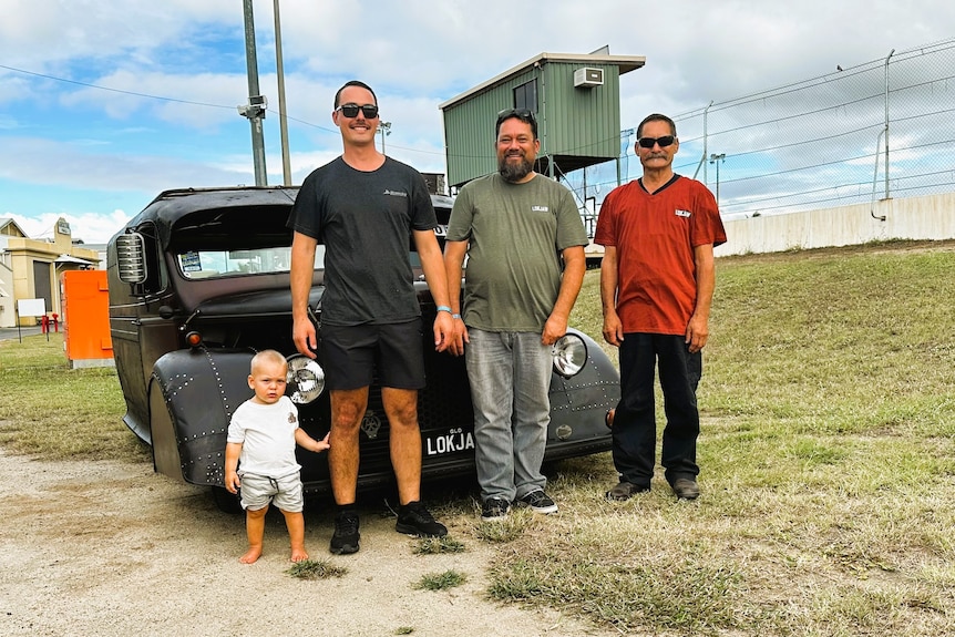 A toddler boy, his dad, granddad and great grandad stand in front of a dark coloured hot rod.