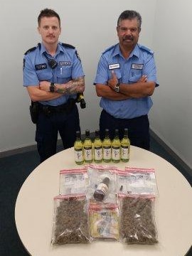 Two WAS police officers stand with their hands crossed behind a table with several evidence bags and bottles of alcohol.