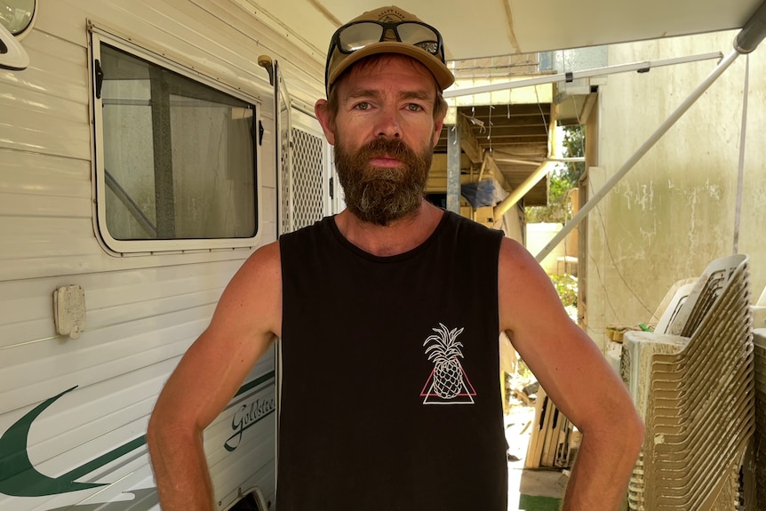 A man in a singlet standing in front of a caravan.