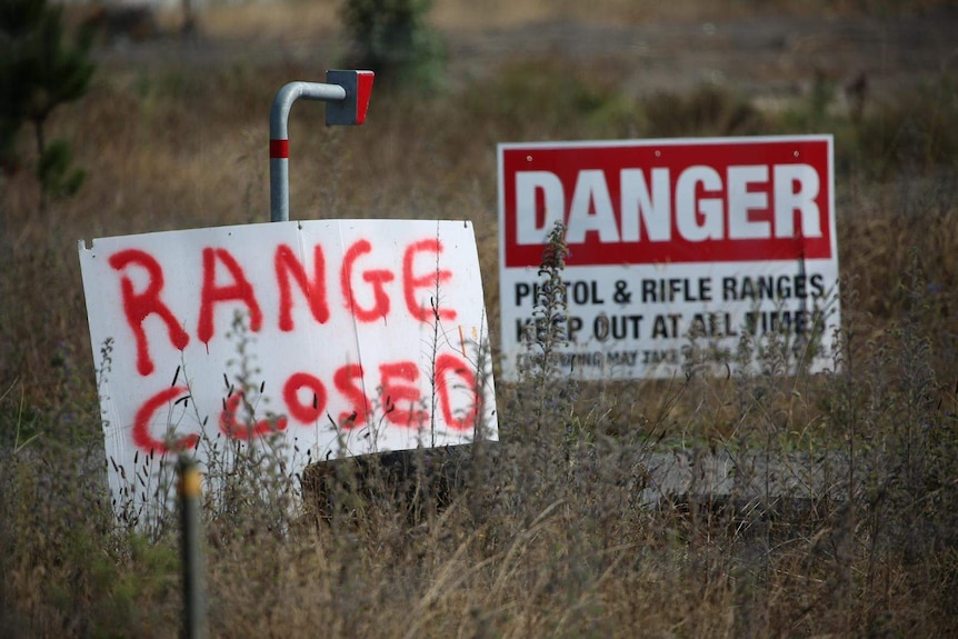 A spray-painted sign that says "range closed" stands in dry grass in front of a warning sign about rifles.