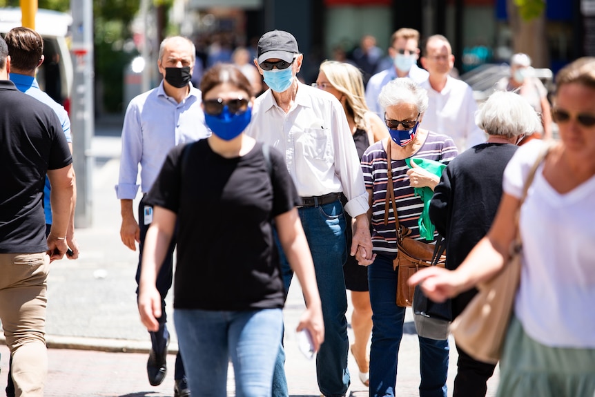 A crowd of people walk across the street in masks, including an older couple.