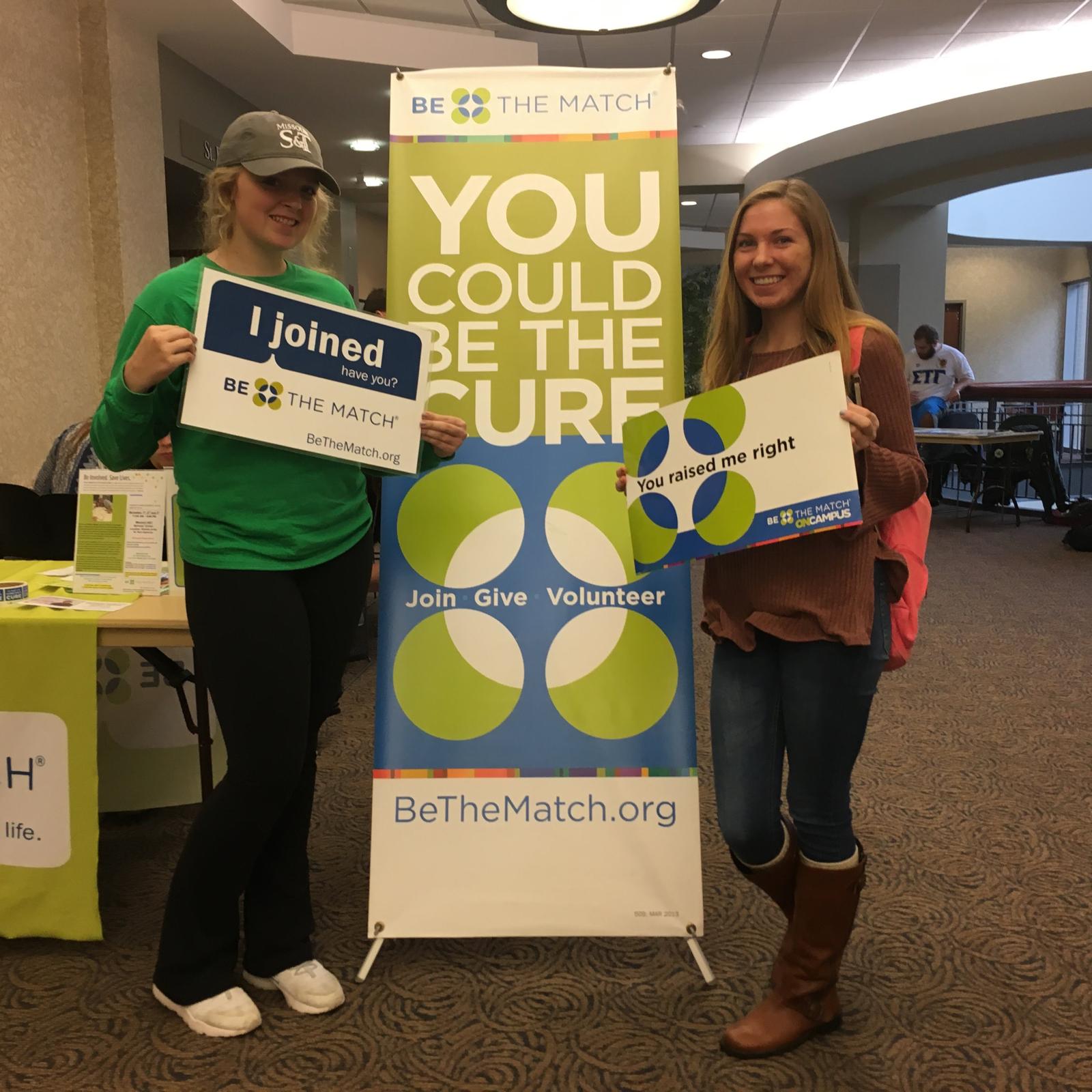 Dos jóvenes rubias sostienen carteles de médula ósea en una sala de conferencias y sonríen a la cámara