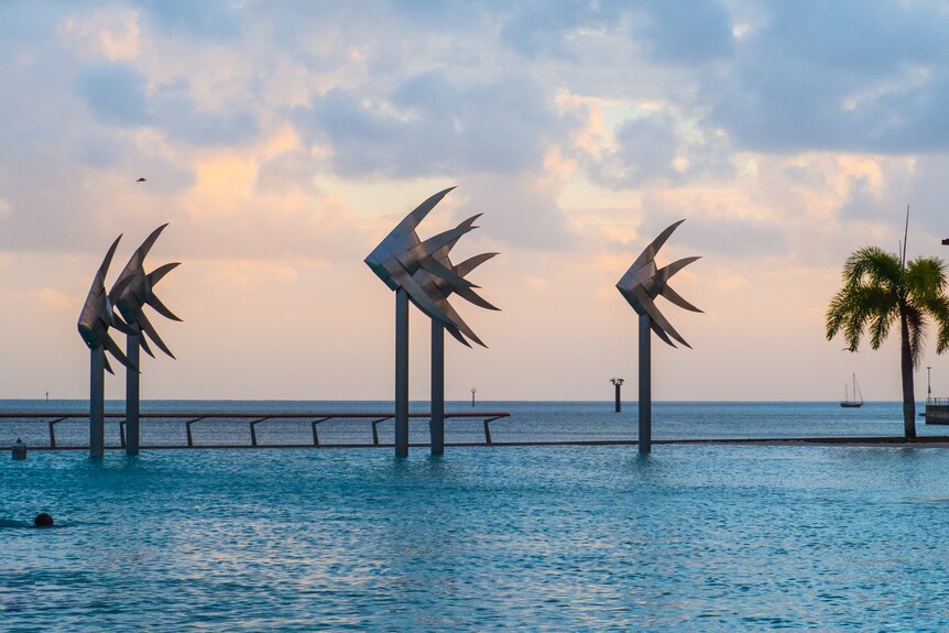 Three steel fish sculptures stand between a swimming pool and the ocean