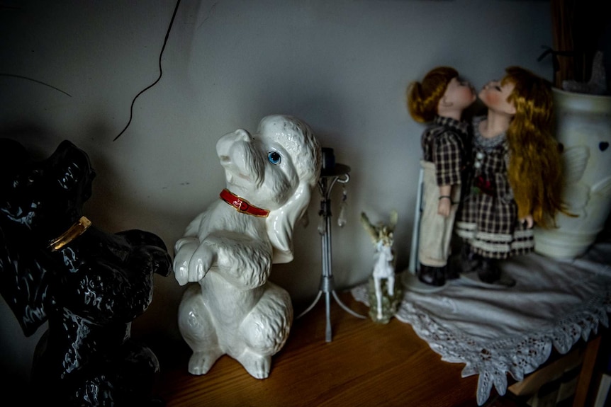Two dog statues stand in the light on a shelf, with two dolls in the background.