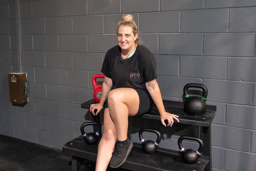 A woman sits on a weights stand holding kettlebells at a gym.