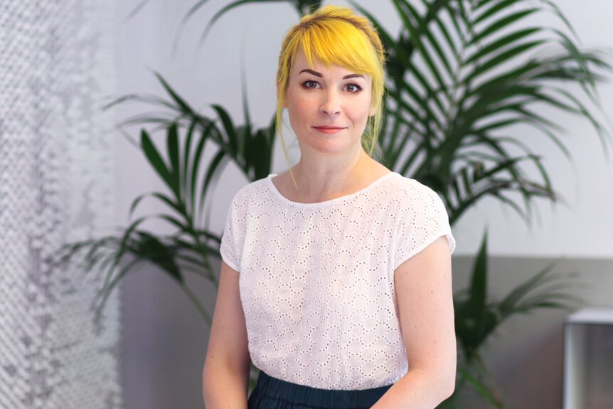 Portrait of a young woman with bright yellow hair sitting on a desk in an office. A large fern can be seen in the background.