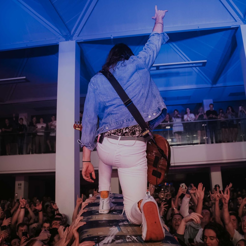 A bass guitarist kneels on a surfboard held up by the crowd.