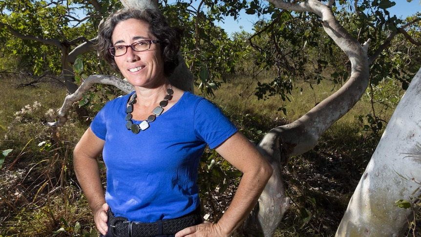 A woman with dark curly hair, wearing glasses and a blue top, standing in front of a gum tree.
