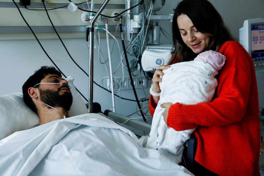 Man lying in hospital bed looks towards his wife standing beside him holding their new born baby.