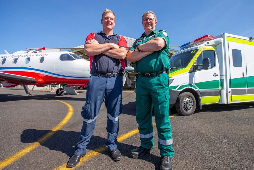 A male RFDS crew member standing next to a male SA Ambulance officer with a plane and ambulance behind them