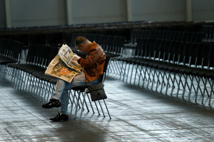An evacuated man reads the paper as he sits alone in a fair trade hall