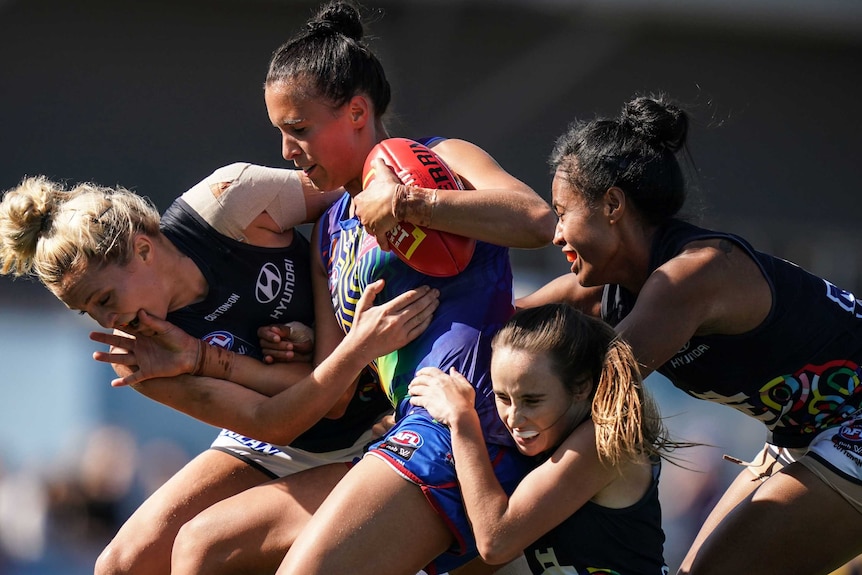 AFLW players converge on a footballer holding ball in her hands - they hang off of her as she runs.