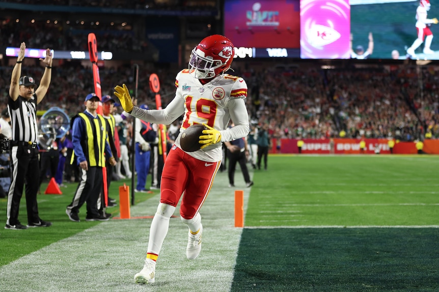 A Kansas City Chiefs player waves to the crowd in the end zone as he holds the ball after scoring a touchdown.