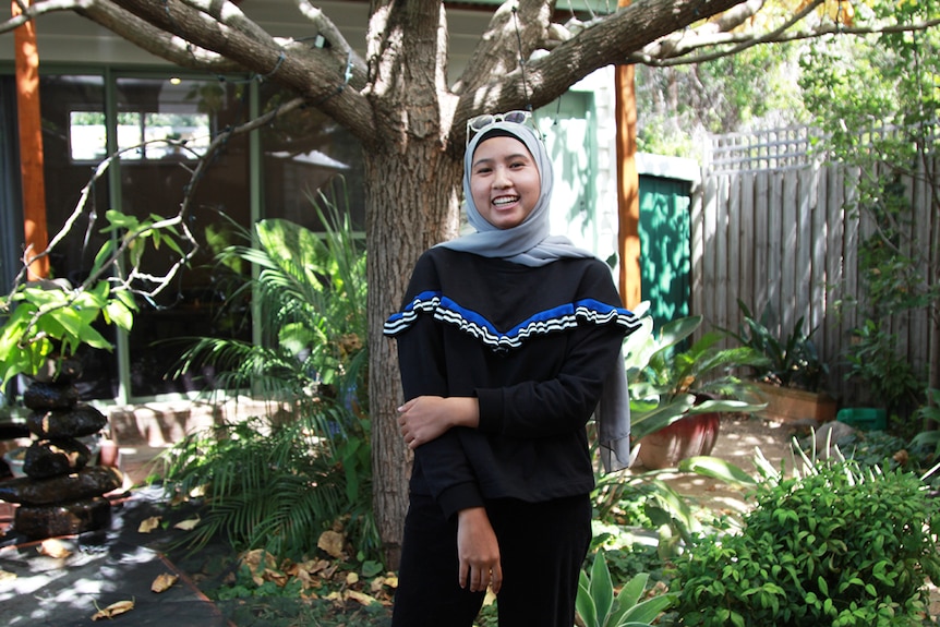 A young woman wearing a light grey hijab and a black jumper stands in a courtyard garden and smiles.