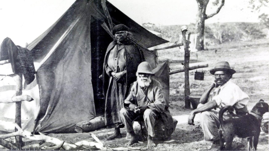 Two men sit on chairs and one woman stands outside a tent.
