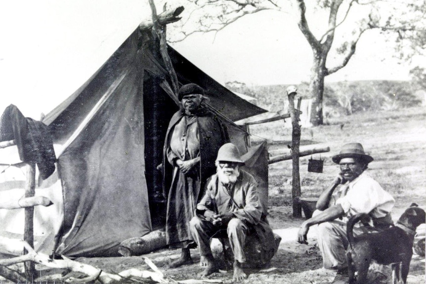 Two men sit on chairs and one woman stands outside a tent.